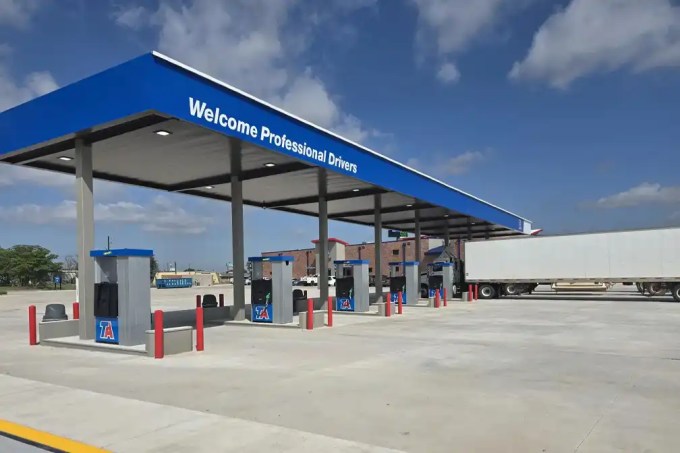 Truck fueling station under a blue canopy that reads 'Welcome Professional Drivers' with several fuel pumps and a parked semi-trailer on the right side in clear weather.