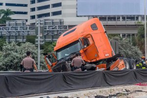 Orange semi truck toppled onto its side on a highway, with responders at the scene and debris nearby.