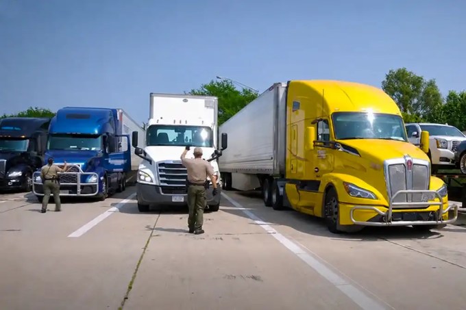 Three large trucks (blue, white, and yellow) parked in a lot with two uniformed officers directing traffic between them—hands raised as signals.