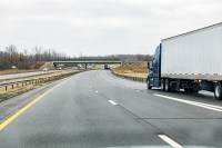 Blue semi-truck driving on a curved multi-lane highway with an overpass in the distance under an overcast sky.